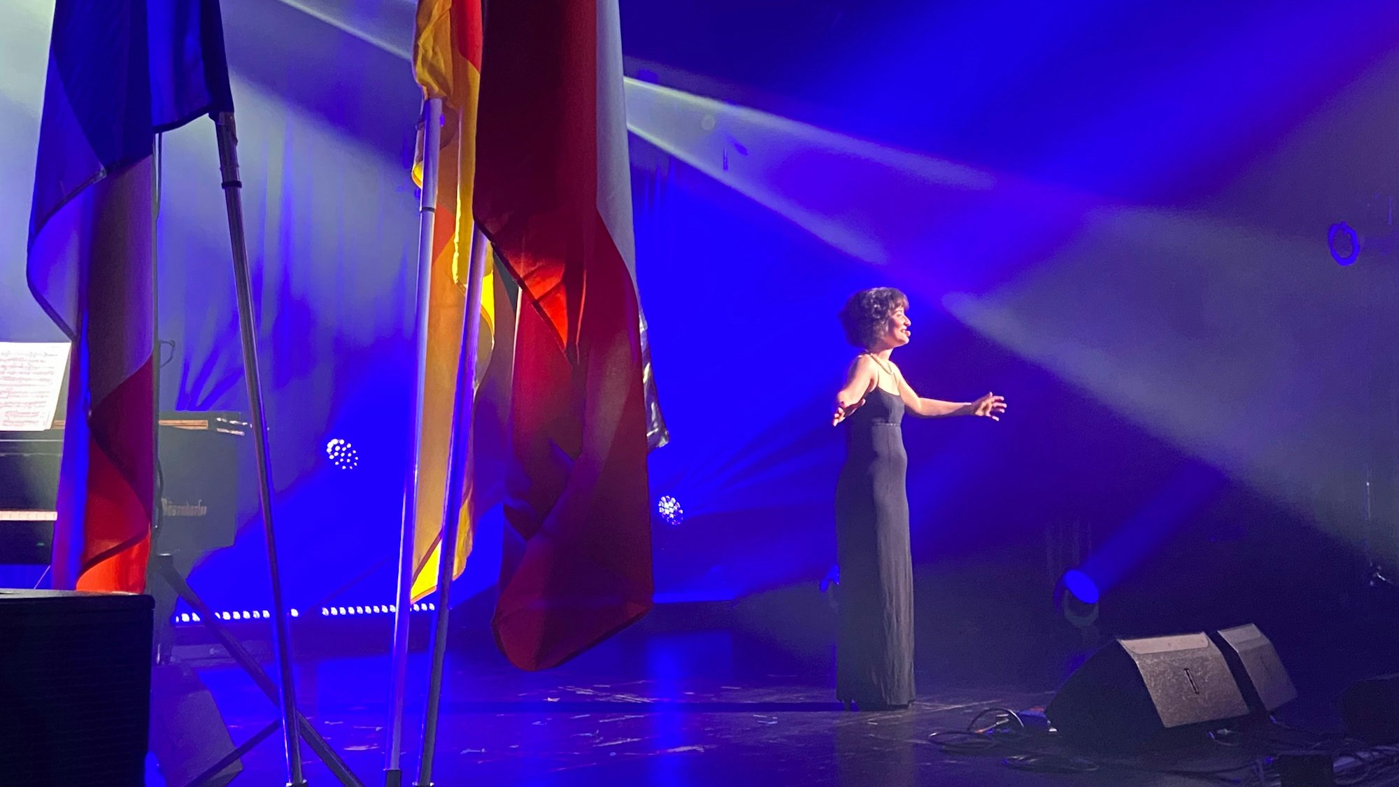 Eine Frau im langen schwarzen Kleid steht mit ausgebreiteten Armen auf einer Bühne.