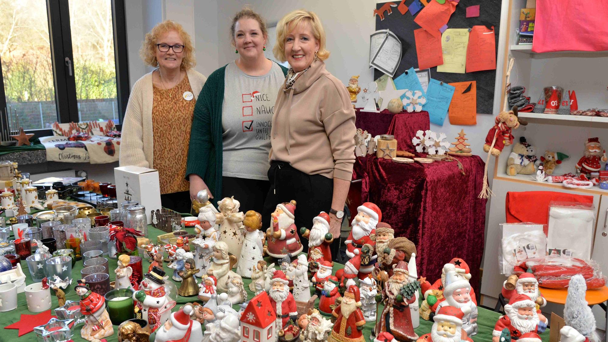 Drei Frauen stehen bei einem Basar an einem Stand, an dem bunte Weihnachtsfiguren verkauft werden.