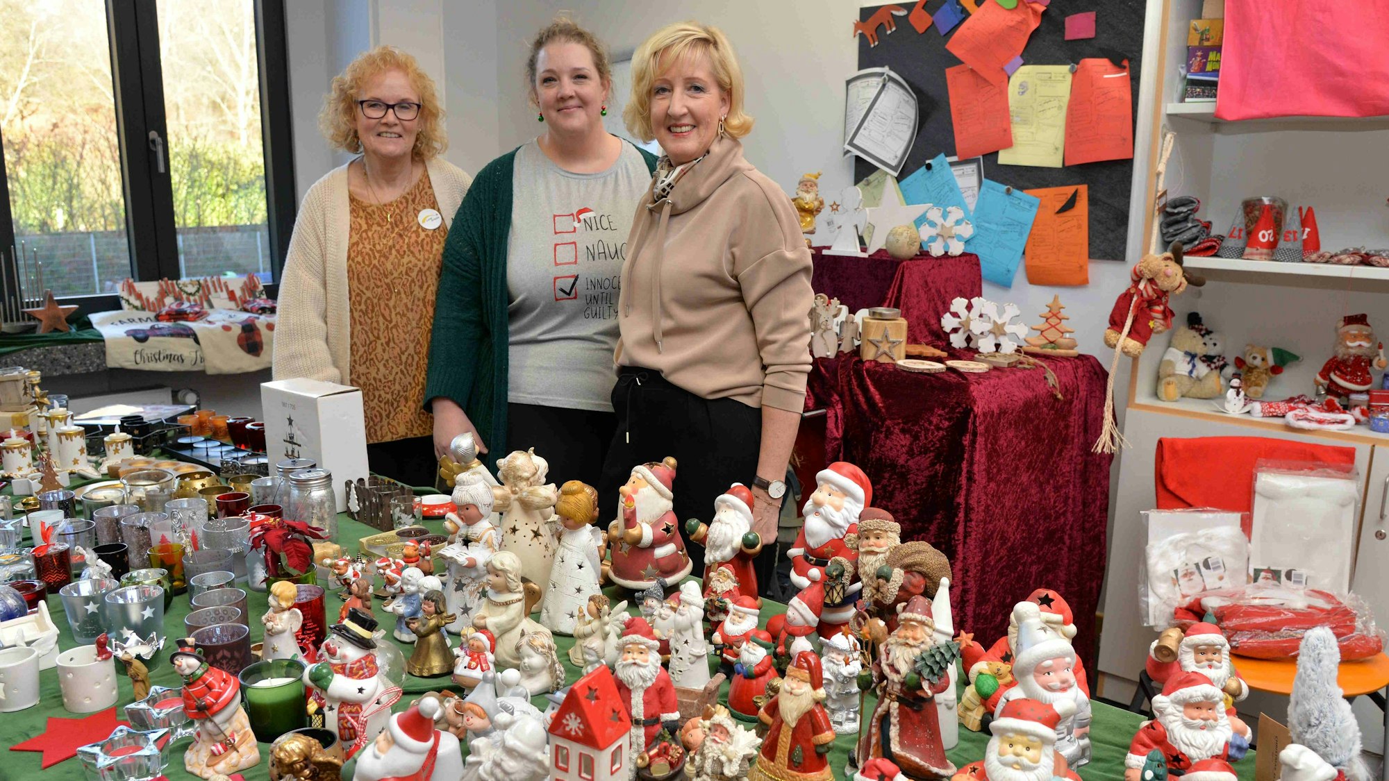 Drei Frauen stehen bei einem Basar an einem Stand, an dem bunte Weihnachtsfiguren verkauft werden.