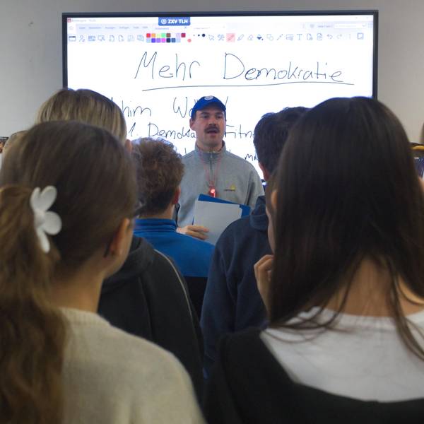 Ein Mann referiert vor einer Schulklasse. Auf der Tafel im Hintergrund steht das Wort „Demokratie“.