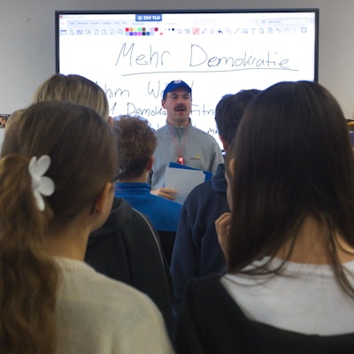 Ein Mann referiert vor einer Schulklasse. Auf der Tafel im Hintergrund steht das Wort „Demokratie“.