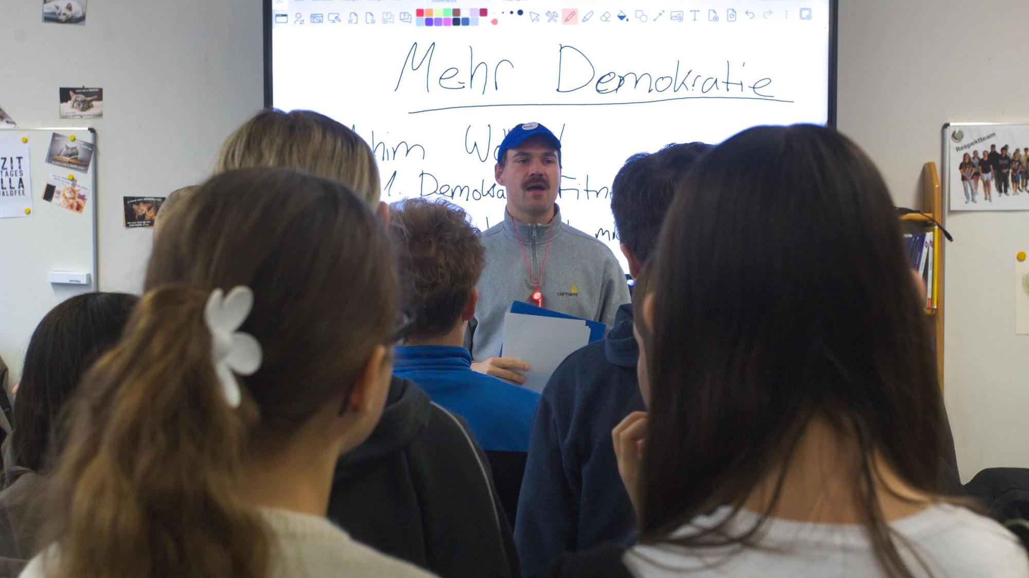 Ein Mann referiert vor einer Schulklasse. Auf der Tafel im Hintergrund steht das Wort „Demokratie“.
