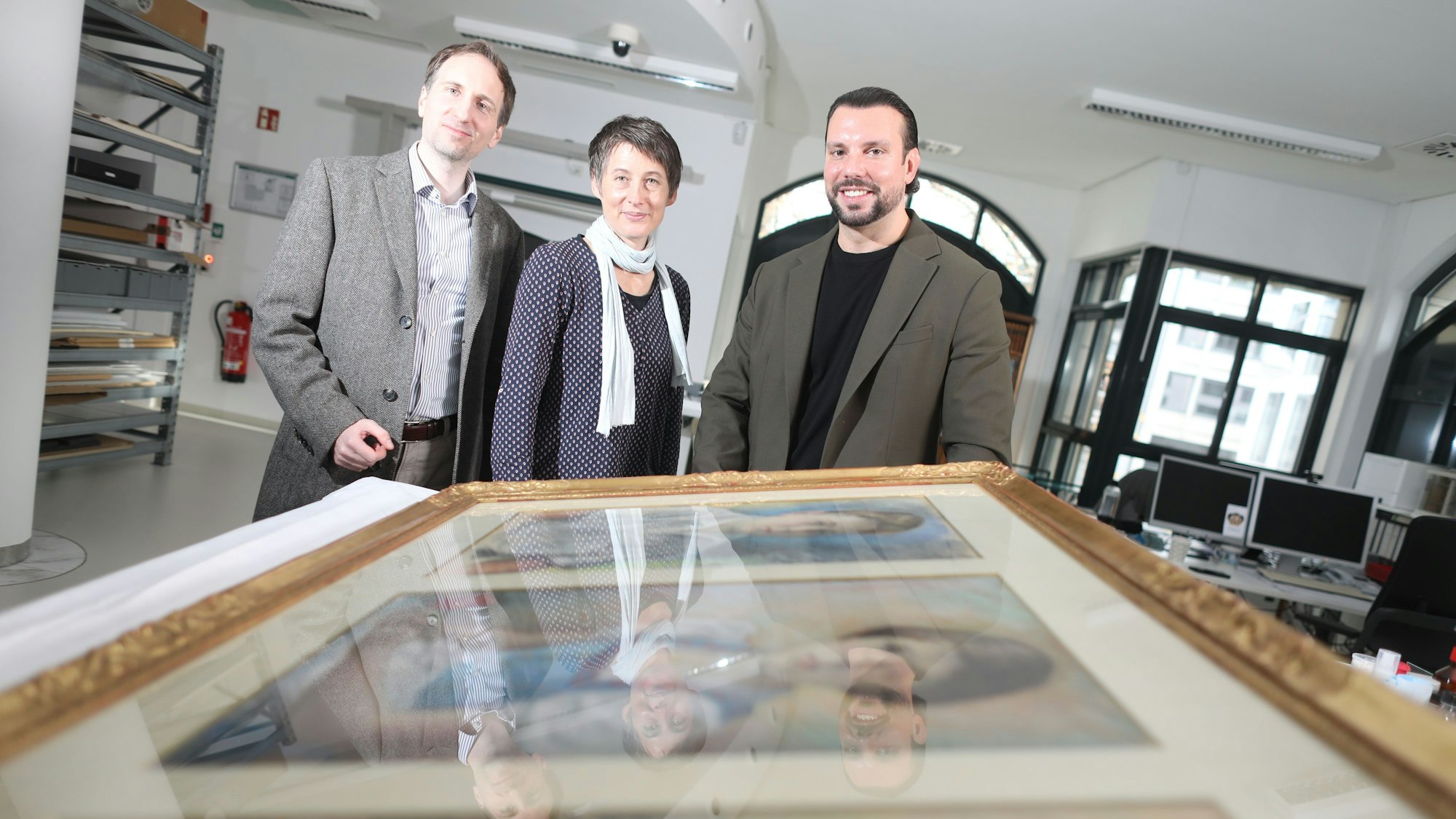 Das Foto zeigt drei Personen vor den eingerahmten Bildern.
