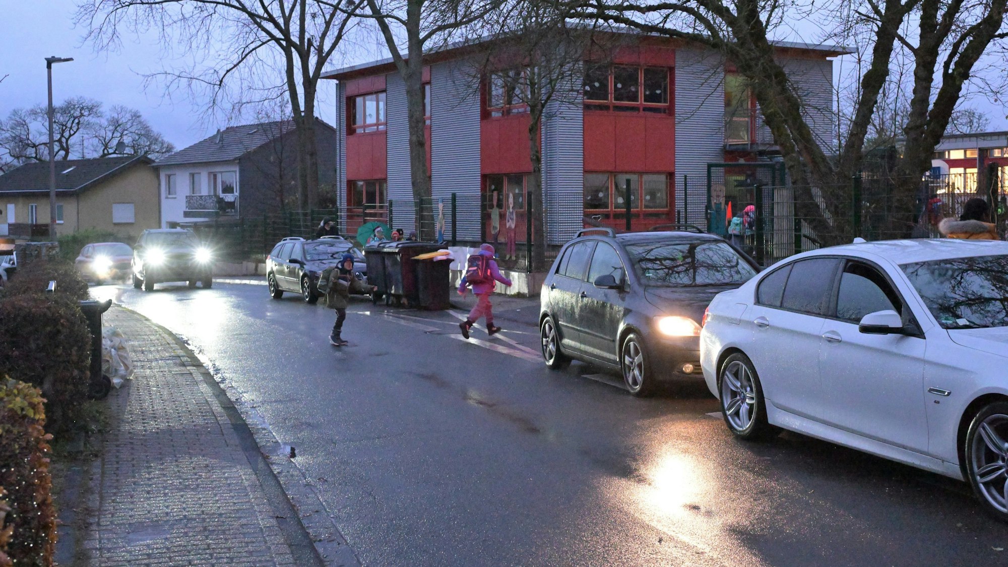 Kinder laufen im Dunkeln über die Straße. Autos kommen angefahren. Fahrzeuge parken am Fahrbahnrand.
