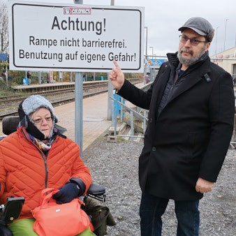 Dulgerakis zeigt mit dem Finger auf ein Schild mit der Aufschrift „Achtung! Rampe nicht barrierefrei. Benutzung auf eigene Gefahr!“