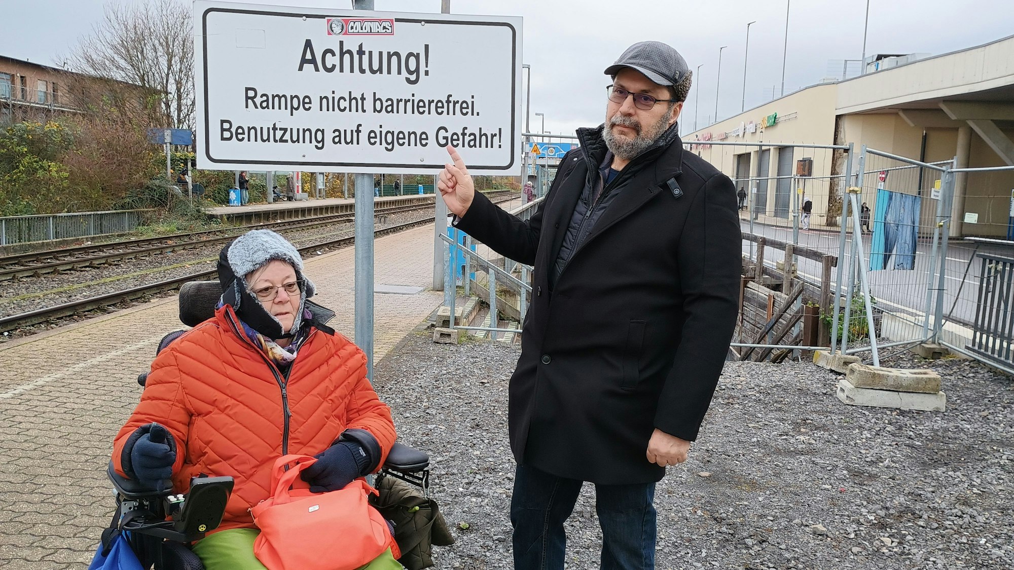 Dulgerakis zeigt mit dem Finger auf ein Schild mit der Aufschrift „Achtung! Rampe nicht barrierefrei. Benutzung auf eigene Gefahr!“