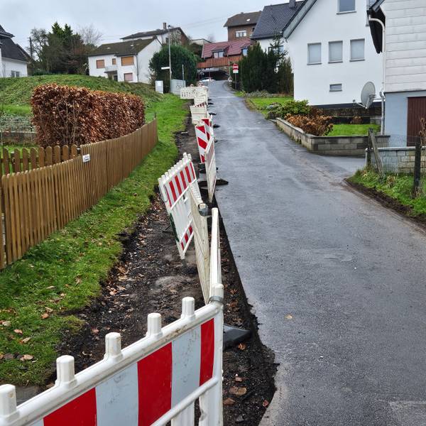 Das Bild zeigt eine Anwohnerstraße mit einer Baustellenabsperrung auf der linken Seite. Dort fehlt die letzte Asphaltschicht.