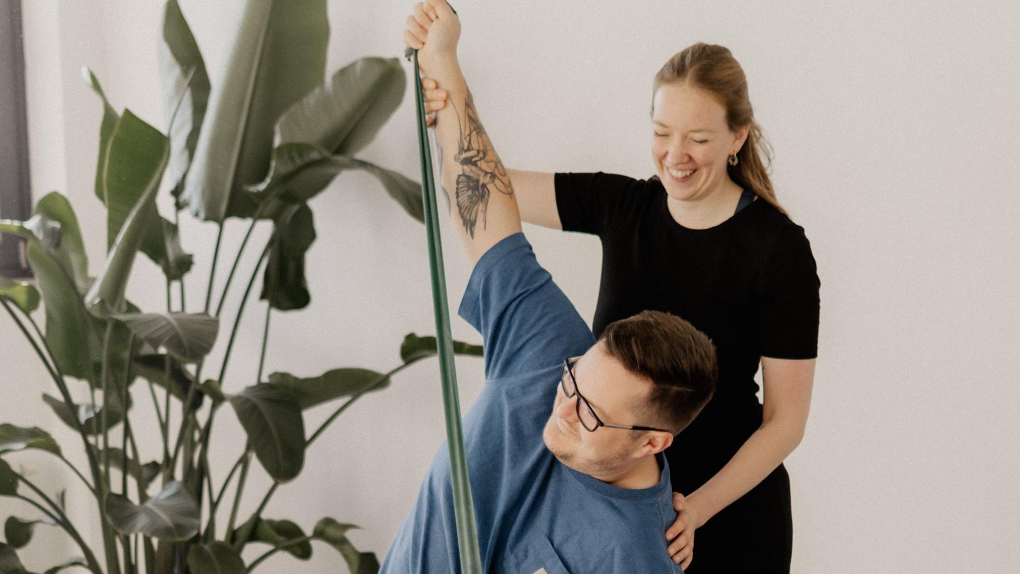 Das Bild zeigt eine Yoga-Lehrerin, die mit einem Mann eine Übung im Sitzen durchführt.