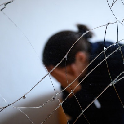 Eine Frau sitzt neben einem gesprungenen Spiegel auf einem Bett und hält ihren Kopf in den Händen.