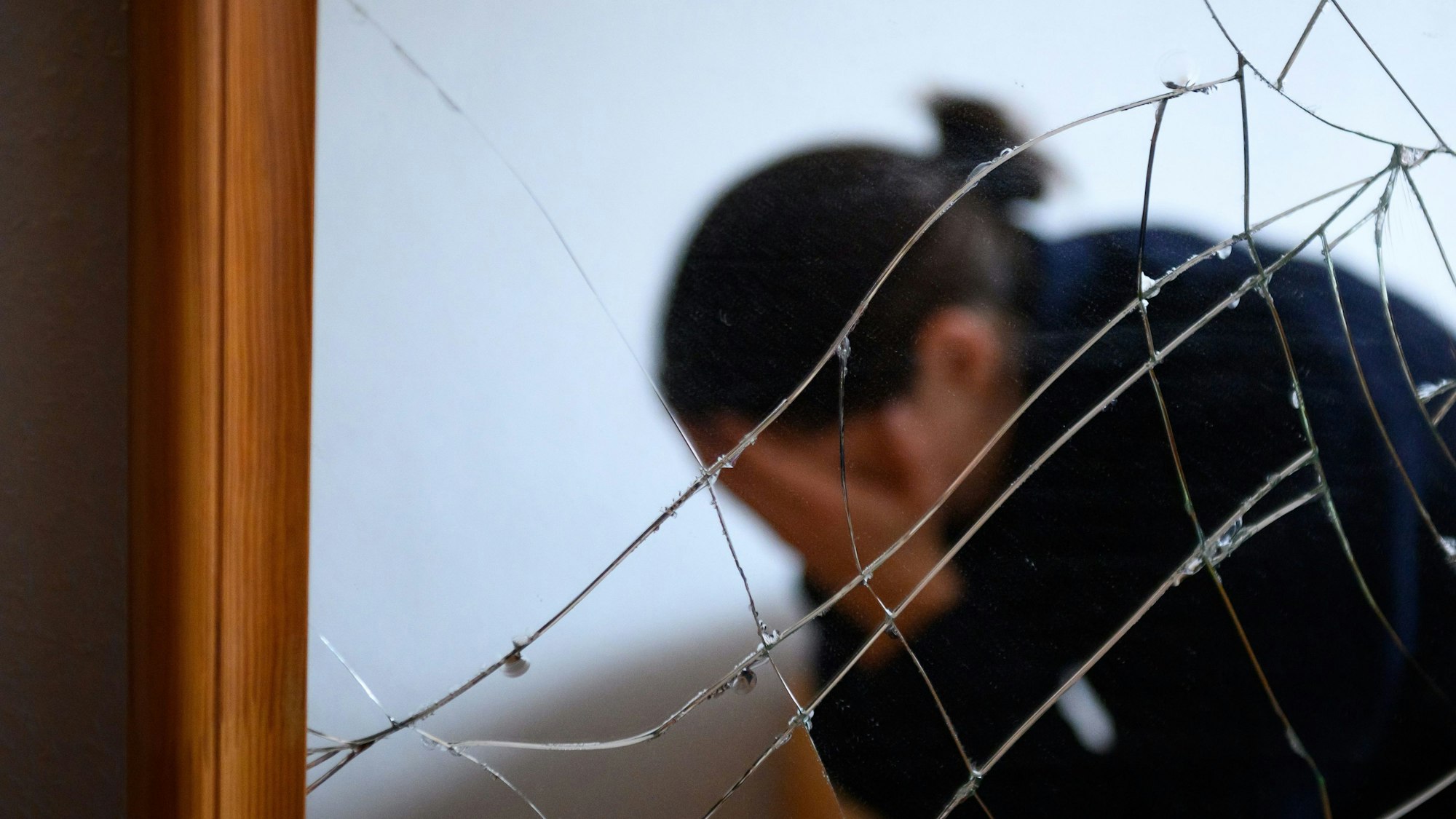 Eine Frau sitzt neben einem gesprungenen Spiegel auf einem Bett und hält ihren Kopf in den Händen (gestellte Szene).