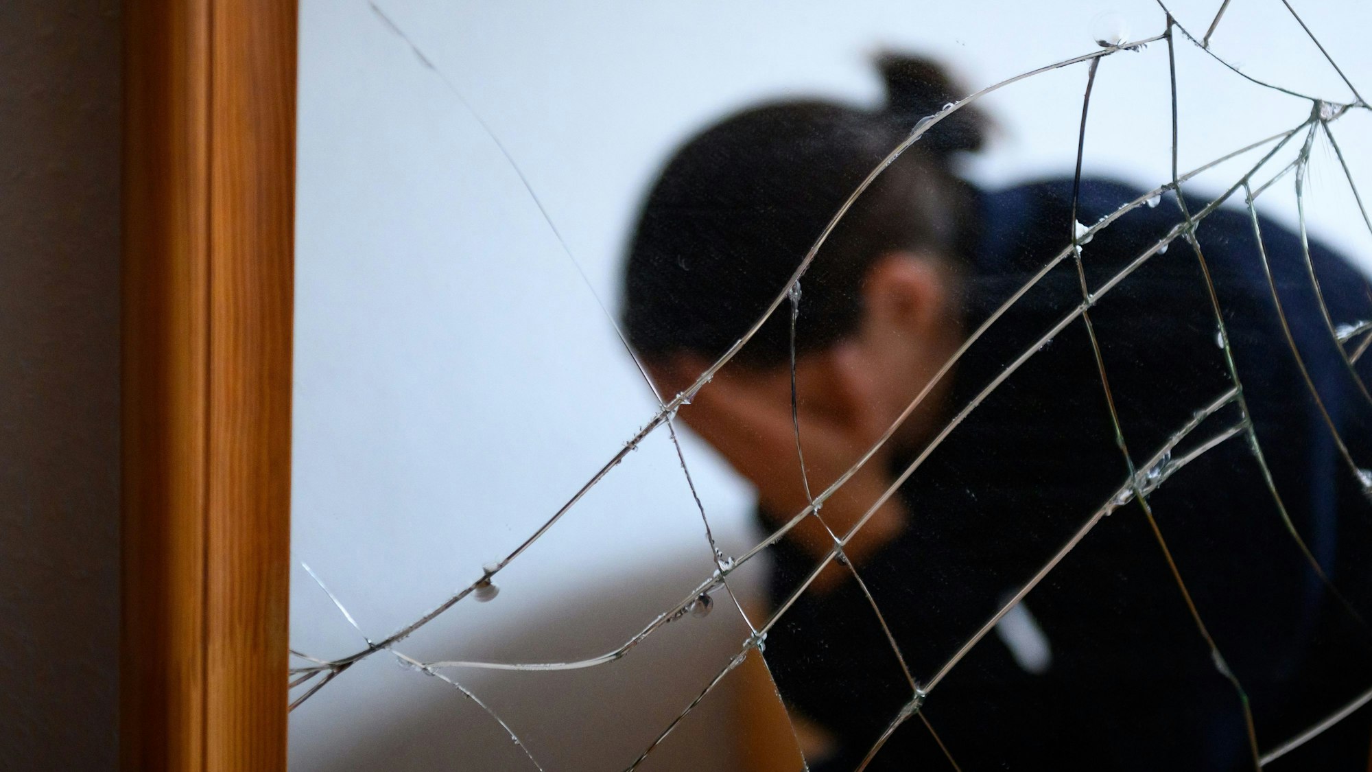 Eine Frau sitzt neben einem gesprungenen Spiegel auf einem Bett und hält ihren Kopf in den Händen.