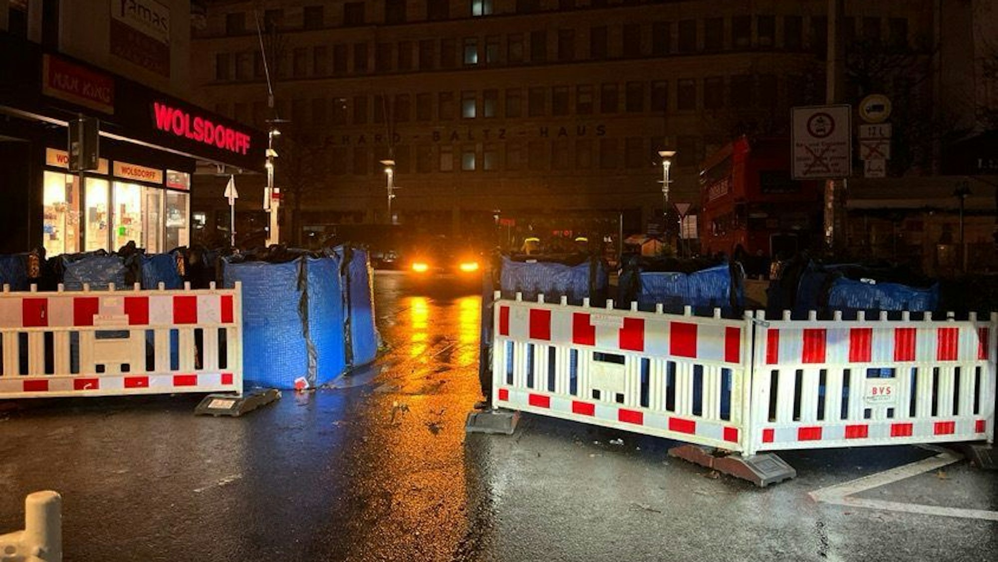Blaue Wassercontainer stehen hinter rot-weißen Absperrbaken auf einer Straße im Dunkeln.