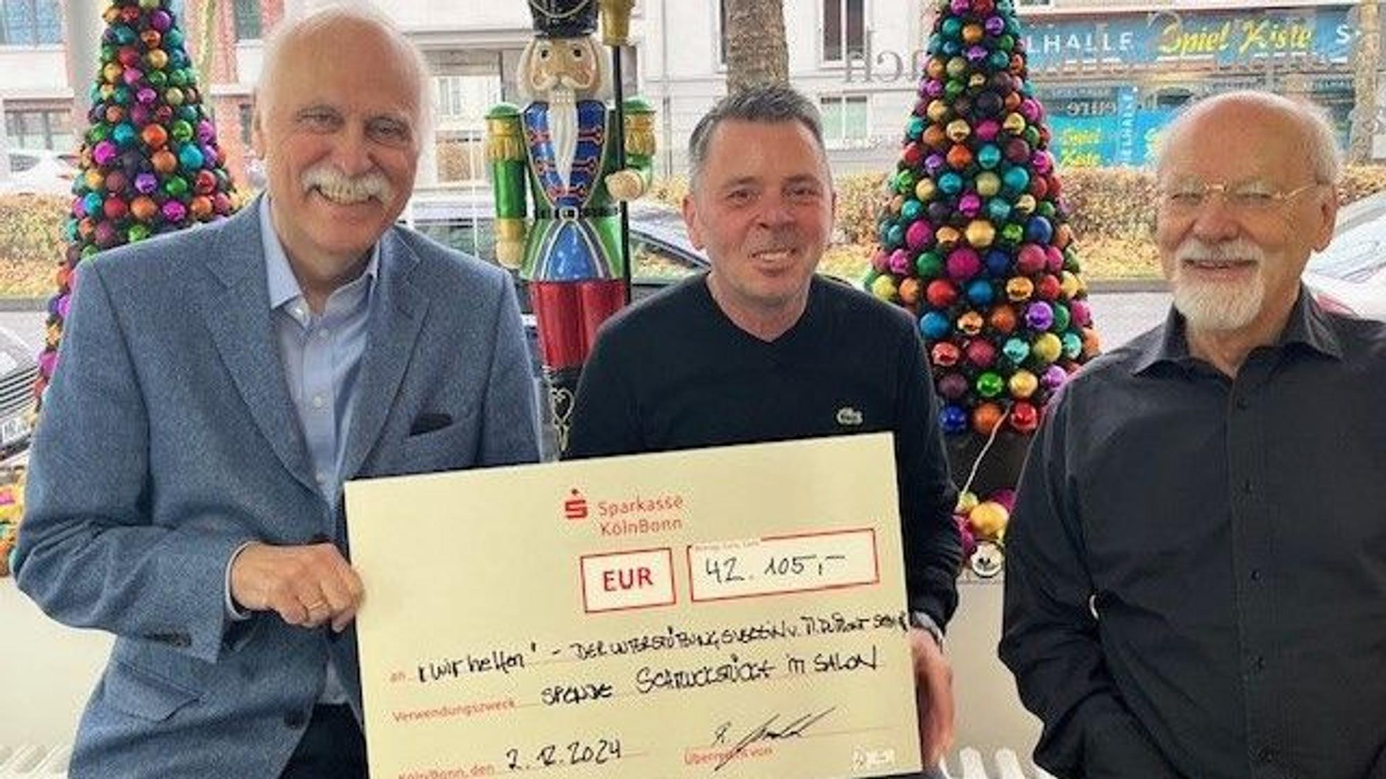 „wir helfen“-Geschäftsführer Karl-Heinz Goßmann, Ralph Schwalbach und Herbert Schmelzer freuen sich über den phänomenalen Spendenrekord in Höhe von 42.105 Euro, sie sitzen vor dem Schaufenster des Klettenberger Salons, in dem bunte Tannenbäume stehen.