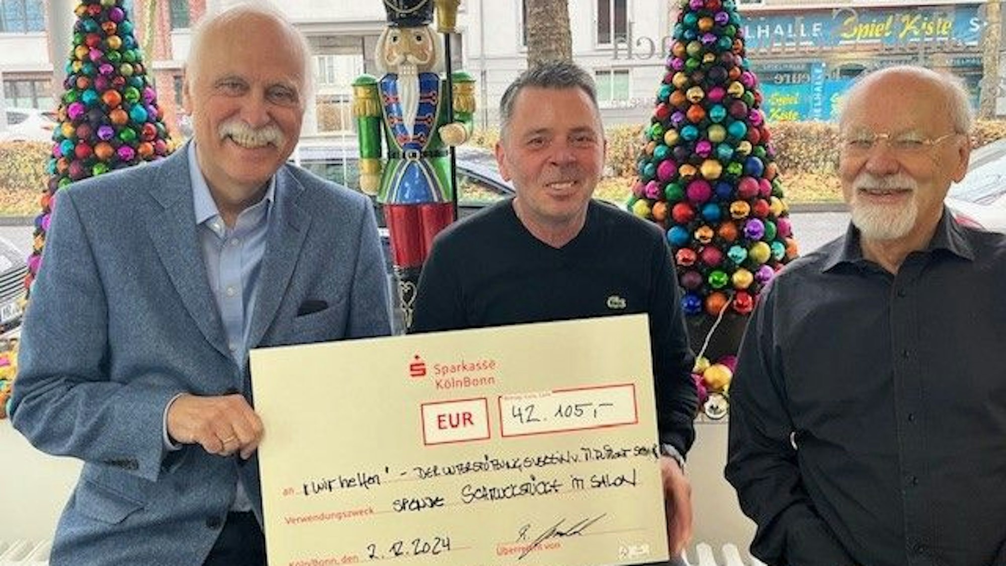 „wir helfen“-Geschäftsführer Karl-Heinz Goßmann, Ralph Schwalbach und Herbert Schmelzer freuen sich über den phänomenalen Spendenrekord in Höhe von 42.105 Euro, sie sitzen vor dem Schaufenster des Klettenberger Salons, in dem bunte Tannenbäume stehen.