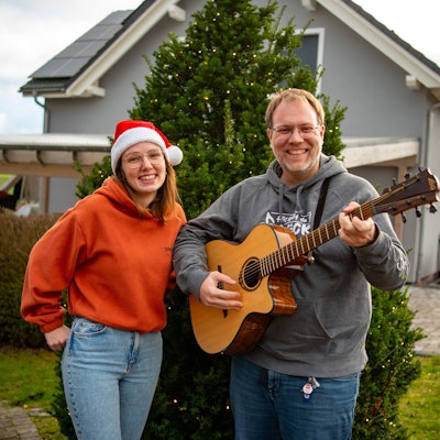 Michael Remer und Anna Jardin von der Sacro-Pop-Band Spirit stehen in Niederbettingen vor einem beleuchteten Tannenbaum. Anna Jardin trägt eine Weihnachtsmütze, einen orangen Pullover und helle Jeans. Michael Renner trägt grauen Pullover und dunkle Jeans und spielt Gitarre.