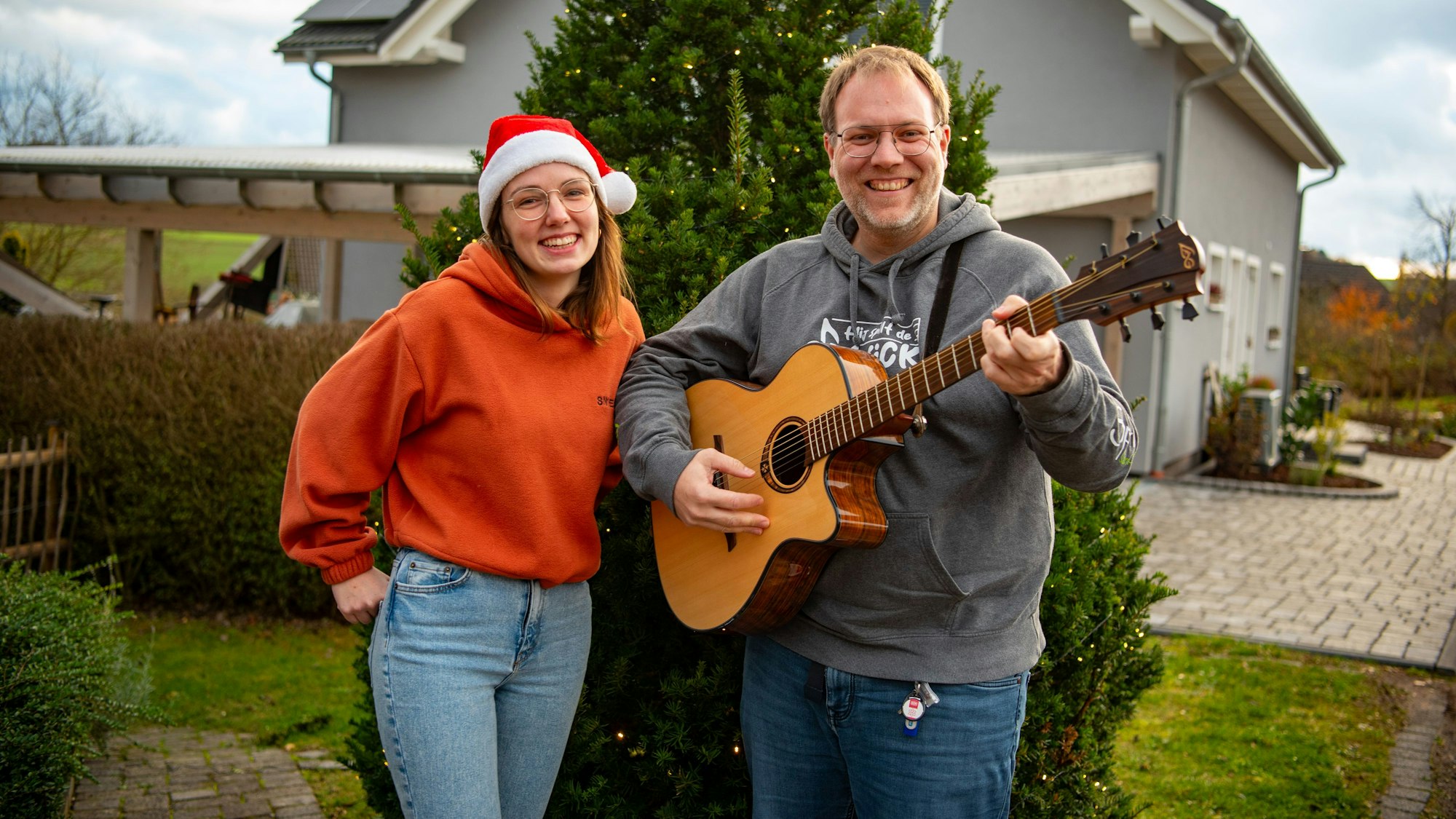 Michael Remer und Anna Jardin von der Sacro-Pop-Band Spirit stehen in Niederbettingen vor einem beleuchteten Tannenbaum. Anna Jardin trägt eine Weihnachtsmütze, einen orangen Pullover und helle Jeans. Michael Renner trägt grauen Pullover und dunkle Jeans und spielt Gitarre.