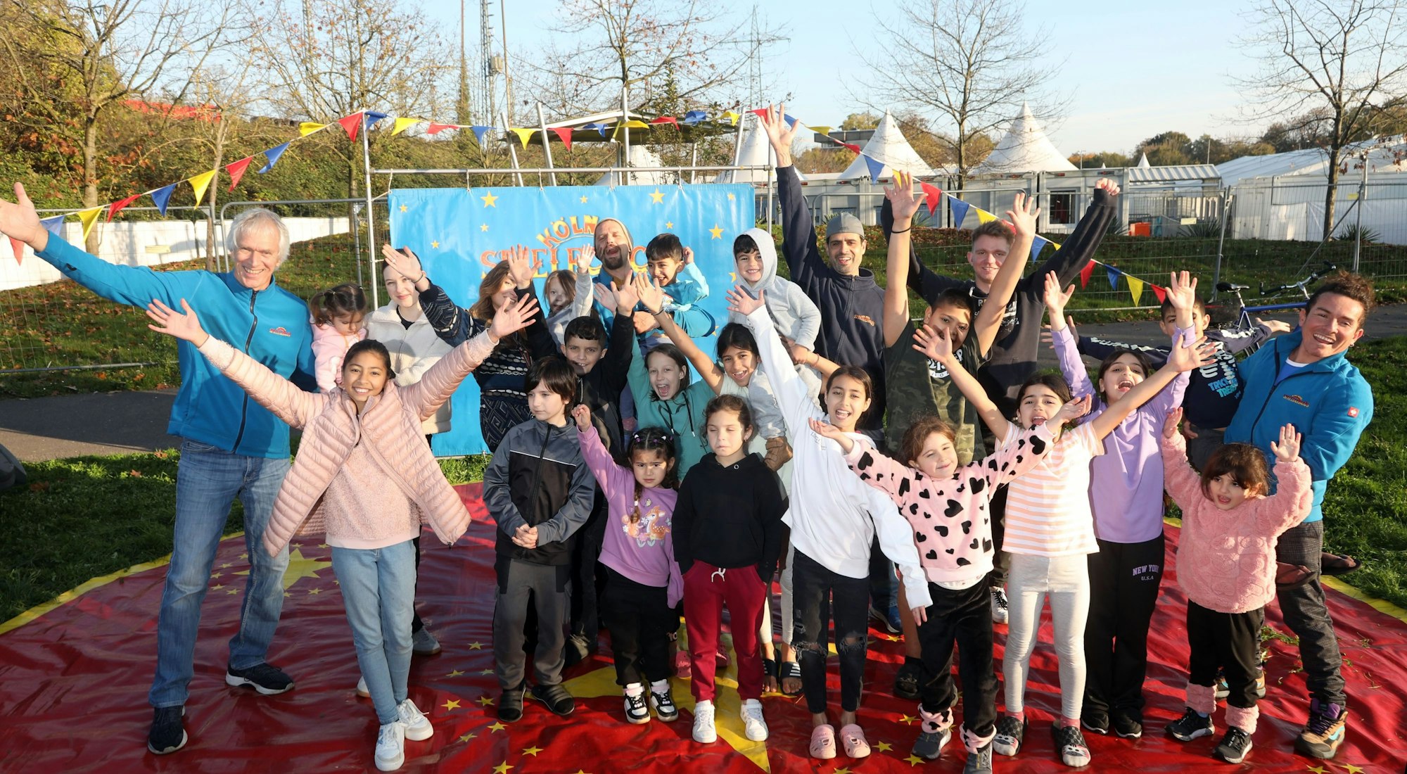 Mitarbeitende des Kölner Spielecircus e.V. und Kinder aus der Notunterkunft für Geflüchtete stehen auf der Manege vor dem Zeltdorf am Südstadion