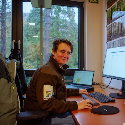 Christa Lang, die Leiterin der Forschungsabteilung im Nationalpark Eifel, sitzt am Schreibtisch in ihrem Büro. Sie trägt eine braune Jacke mit dem National-Park-Logo auf dem Ärmel. Hinter ihrem Bildschirm hängen Fotos und Karten vom Nationalpark, durch ein Fenster sieht man Wald.