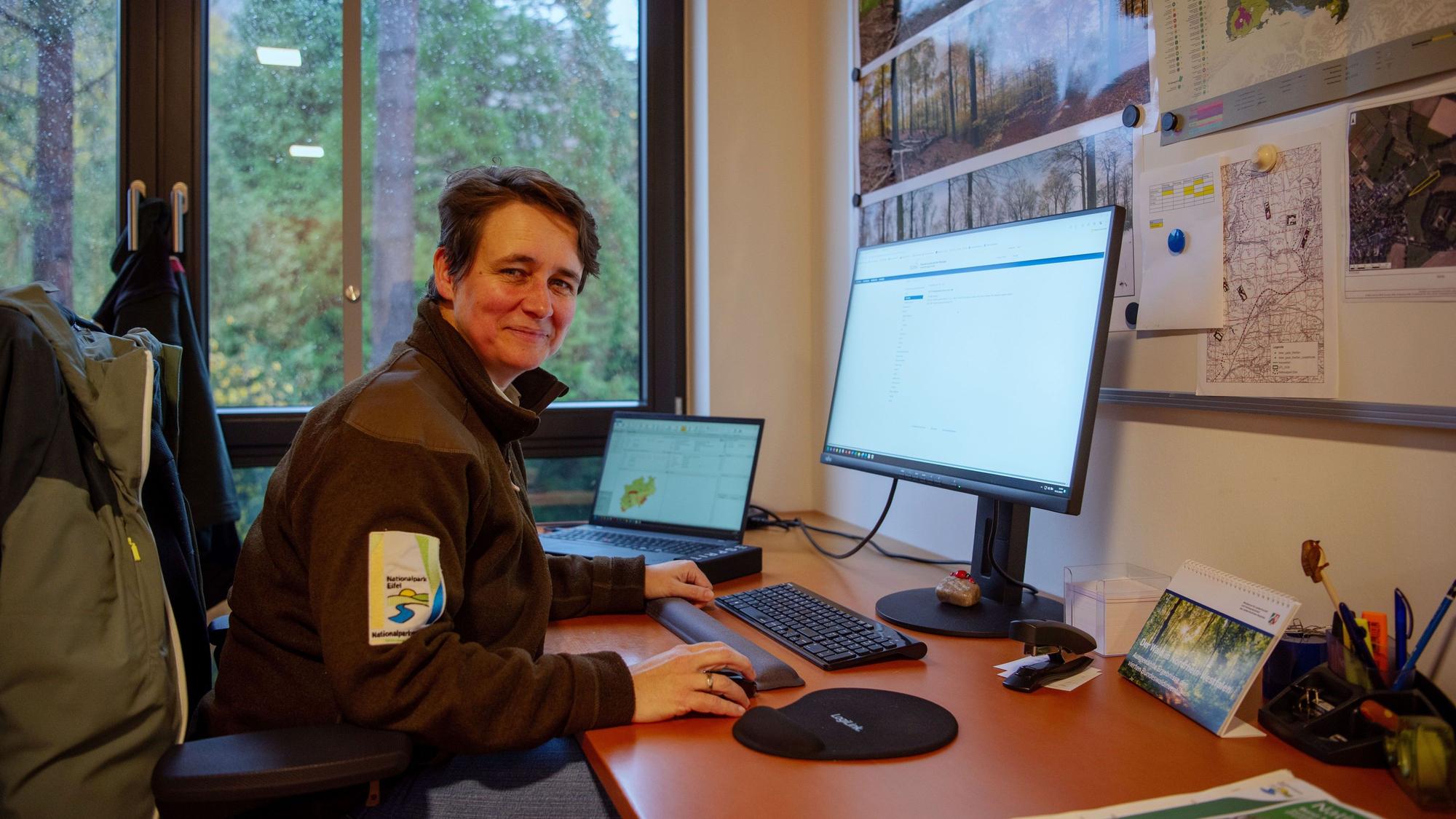 Christa Lang, die Leiterin der Forschungsabteilung im Nationalpark Eifel, sitzt am Schreibtisch in ihrem Büro. Sie trägt eine braune Jacke mit dem National-Park-Logo auf dem Ärmel. Hinter ihrem Bildschirm hängen Fotos und Karten vom Nationalpark, durch ein Fenster sieht man Wald.