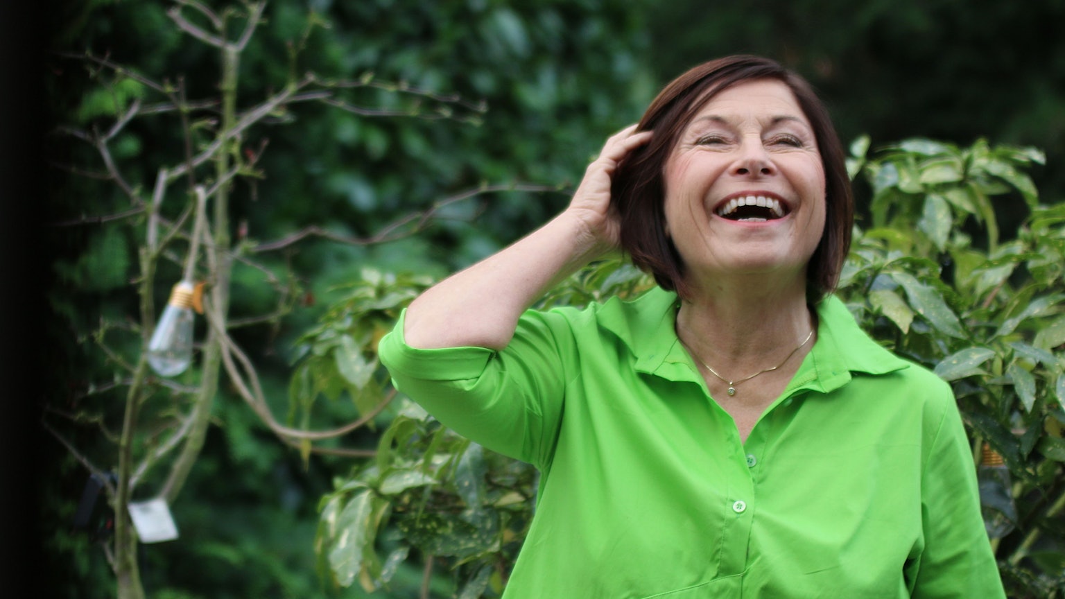 Brigitte Schröder steht in ihrem Garten. Sie lacht und wischt sich mit der Hand ihre Haare aus dem Gesicht.
