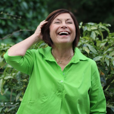 Brigitte Schröder steht in ihrem Garten. Sie lacht und wischt sich mit der Hand ihre Haare aus dem Gesicht.