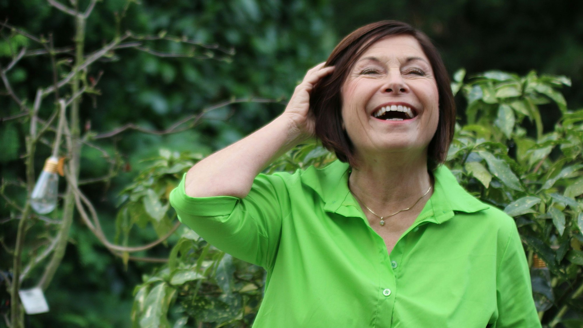 Brigitte Schröder steht in ihrem Garten. Sie lacht und wischt sich mit der Hand ihre Haare aus dem Gesicht.