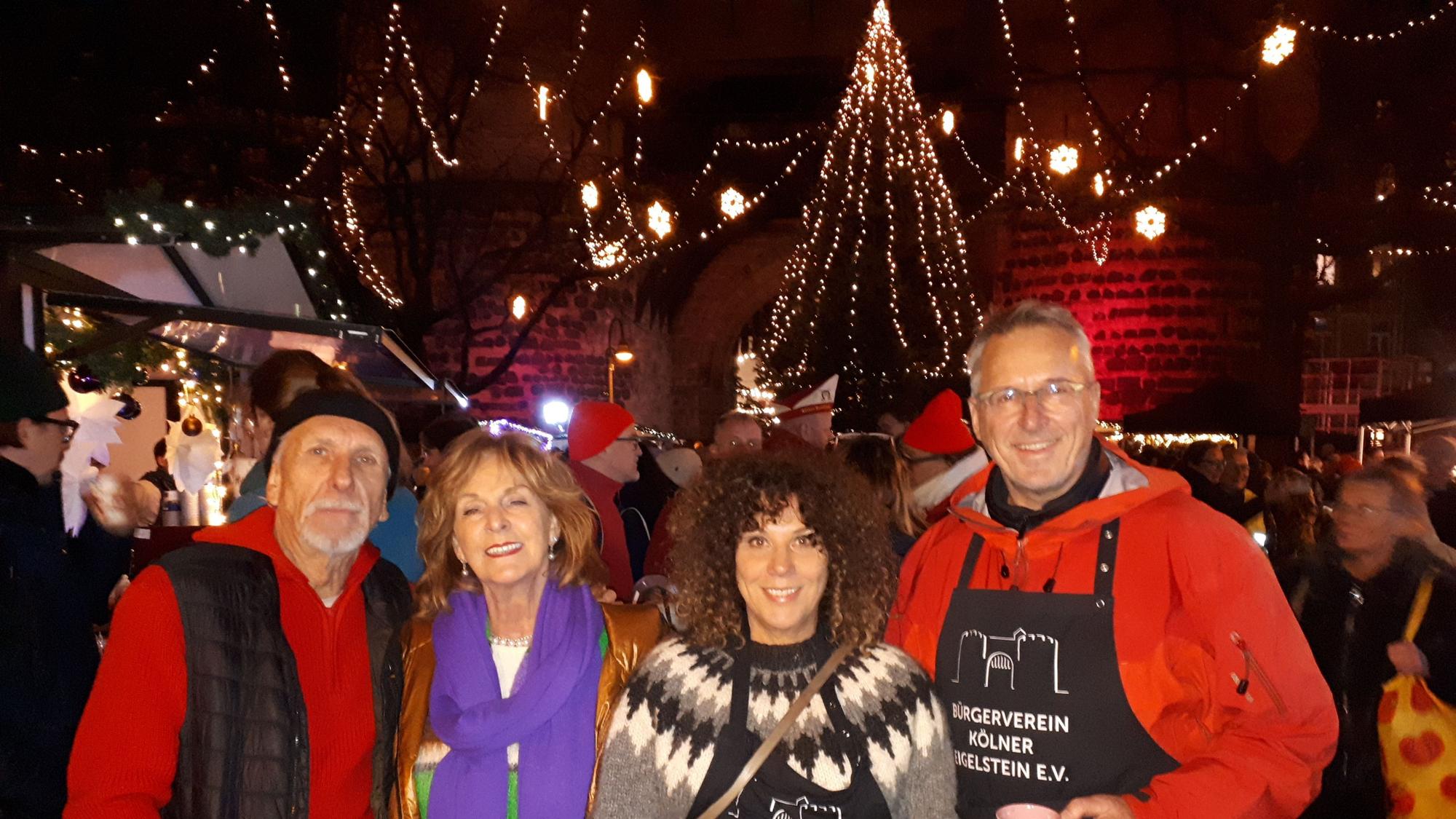 Vier Menschen stehen abends vor der festlich geschmückten Eigelsteintorburg. Im Hintergrund steht auch ein beleuchteter großer Weihnachtsbaum.