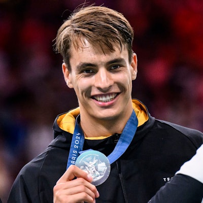 Handballer Julian Köster zeigt lächelnd seine olympische Silbermedaille in die Kamera.