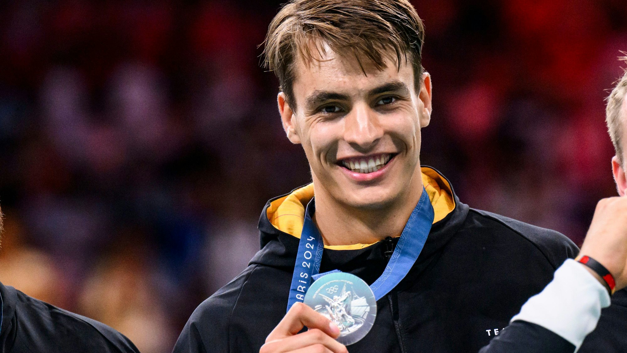 Handballer Julian Köster zeigt lächelnd seine olympische Silbermedaille in die Kamera.