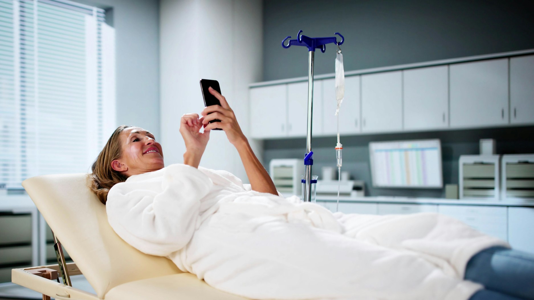 Eine Frau liegt lächelnd auf einer Liege in einem Behandlungszimmer und schaut auf ihr Handy. Nebenbei erhält sie eine Infusion