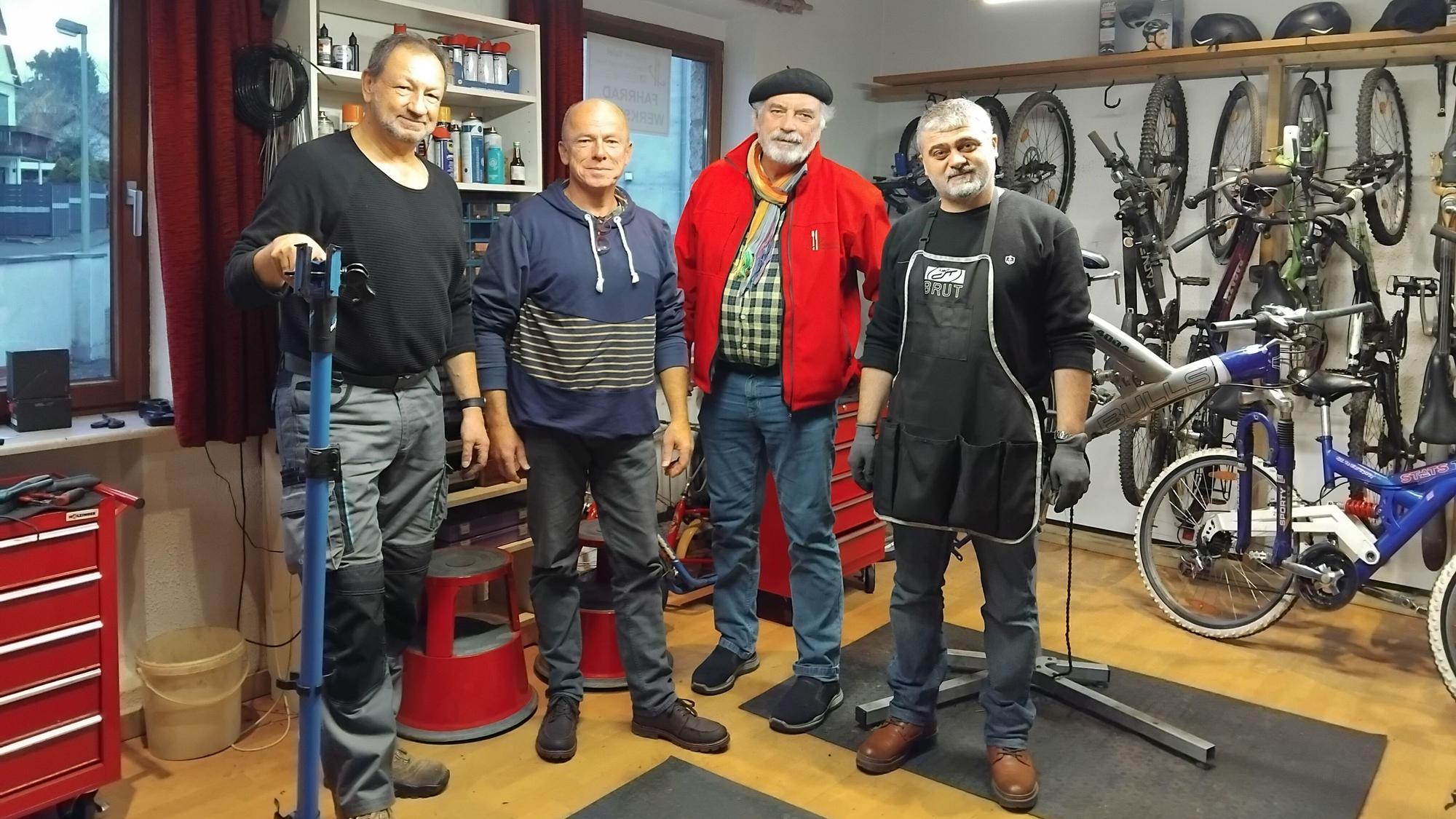 Vier Männer stehen in einer Werkstatt, an der Wand sieht man mehrere Fahrräder.
