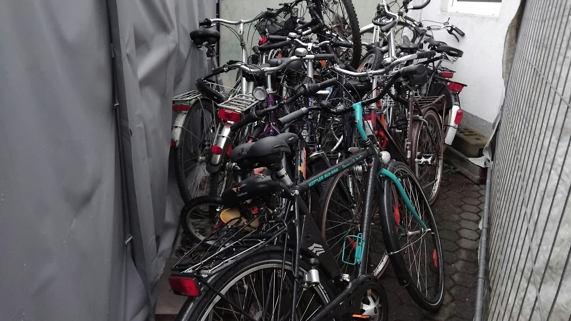 Mehrere Fahrräder stehen zwischen einer Plane und einem Gitter in einem Innenhof und sind nass vom Regen.