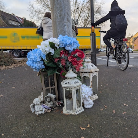 Das Foto zeigt die kleine Gedenkstätte vor dem Ampelmast an der Kreuzung, im Hintergrund ein Radfahrer und Fußgänger.
