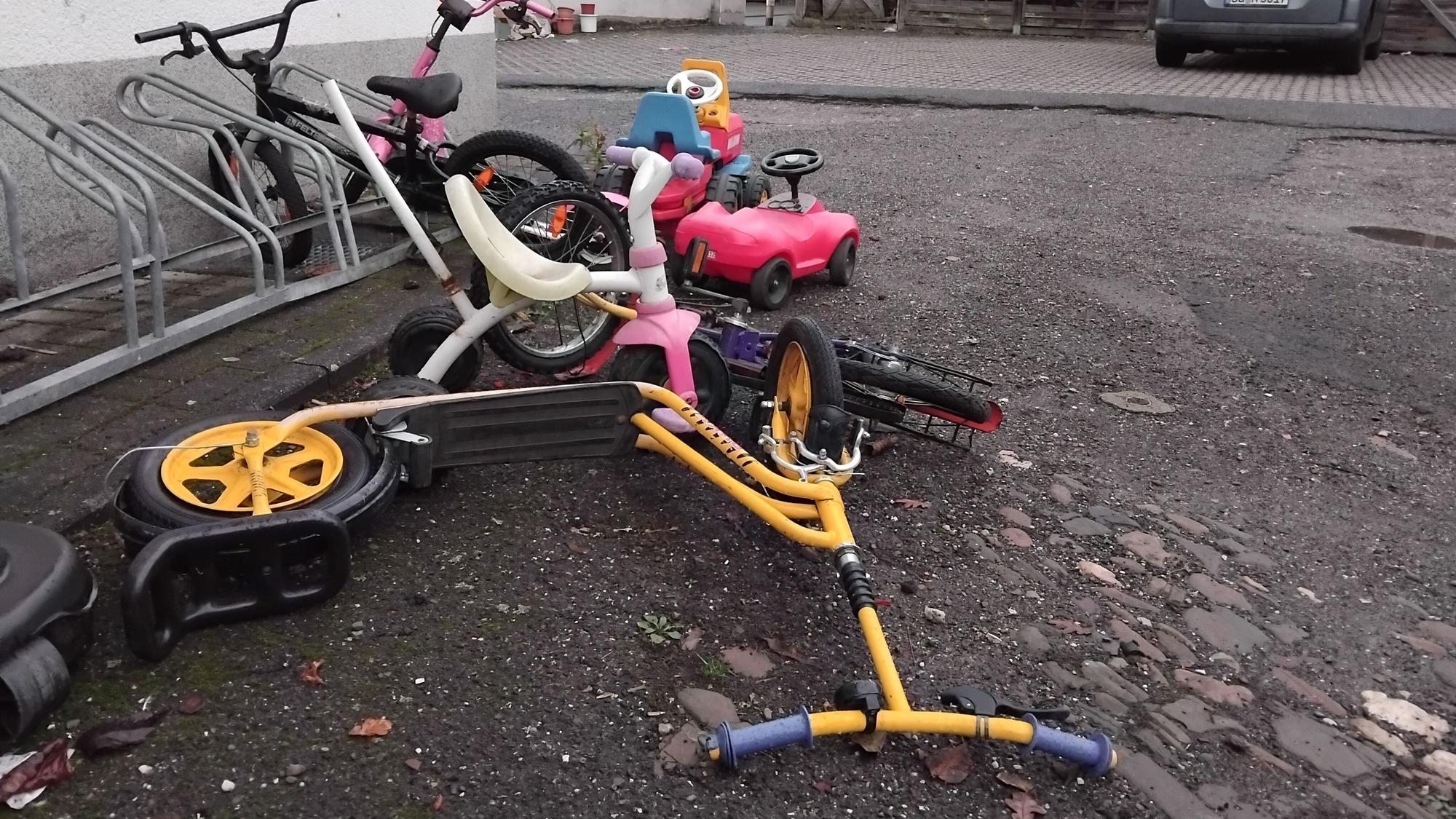 Ein paar bunte Kinderräder, Kickroller und ein Bobbycar liegen in einem Hof.