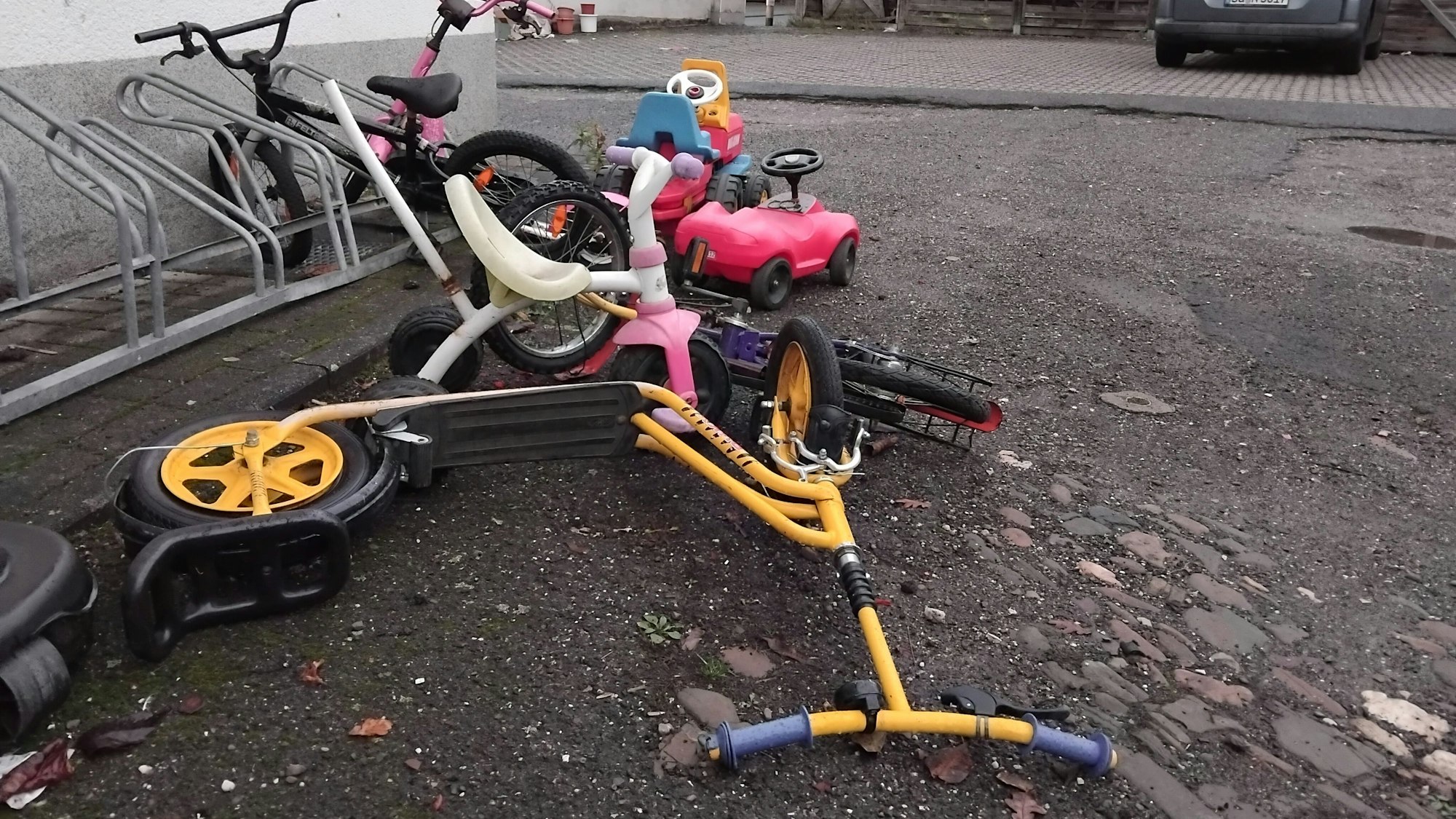 Ein paar bunte Kinderräder, Kickroller und ein Bobbycar liegen in einem Hof.