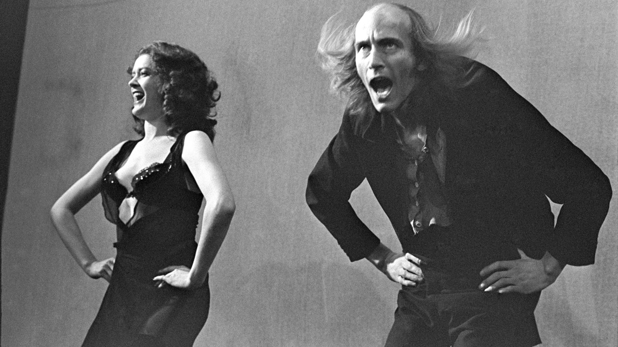 Schwarz-Weiß-Foto von der Premiere der „Rocky Horror Show“ im Royal Court Theatre Upstairs von Douglas H Jeffery: Patricia Quinn trägt als Magenta ein offenes Kleid und einen schwarzen BH, Richard O'Brien als Riff Raff ein schwarzes Jackett.