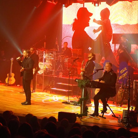 Die Musikgruppe Höhner steht in Schwarz gekleidet auf einer in rotes Licht getauchten Bühne. Im Hintergrund ist ein Foto eines Menschen, der ein Baby hochhält, eingeblendet.