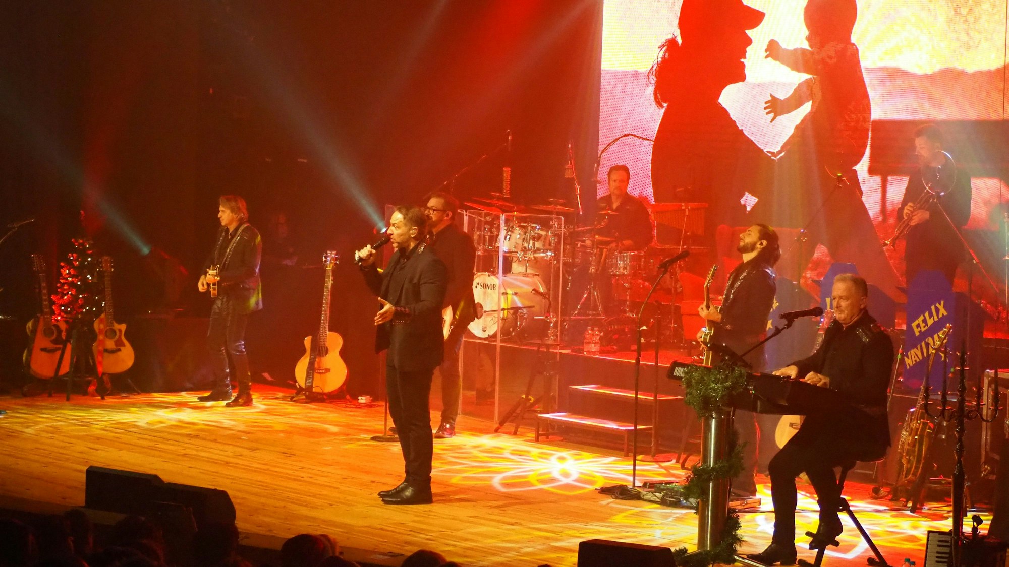 Die Musikgruppe Höhner steht in Schwarz gekleidet auf einer in rotes Licht getauchten Bühne. Im Hintergrund ist ein Foto eines Menschen, der ein Baby hochhält, eingeblendet.