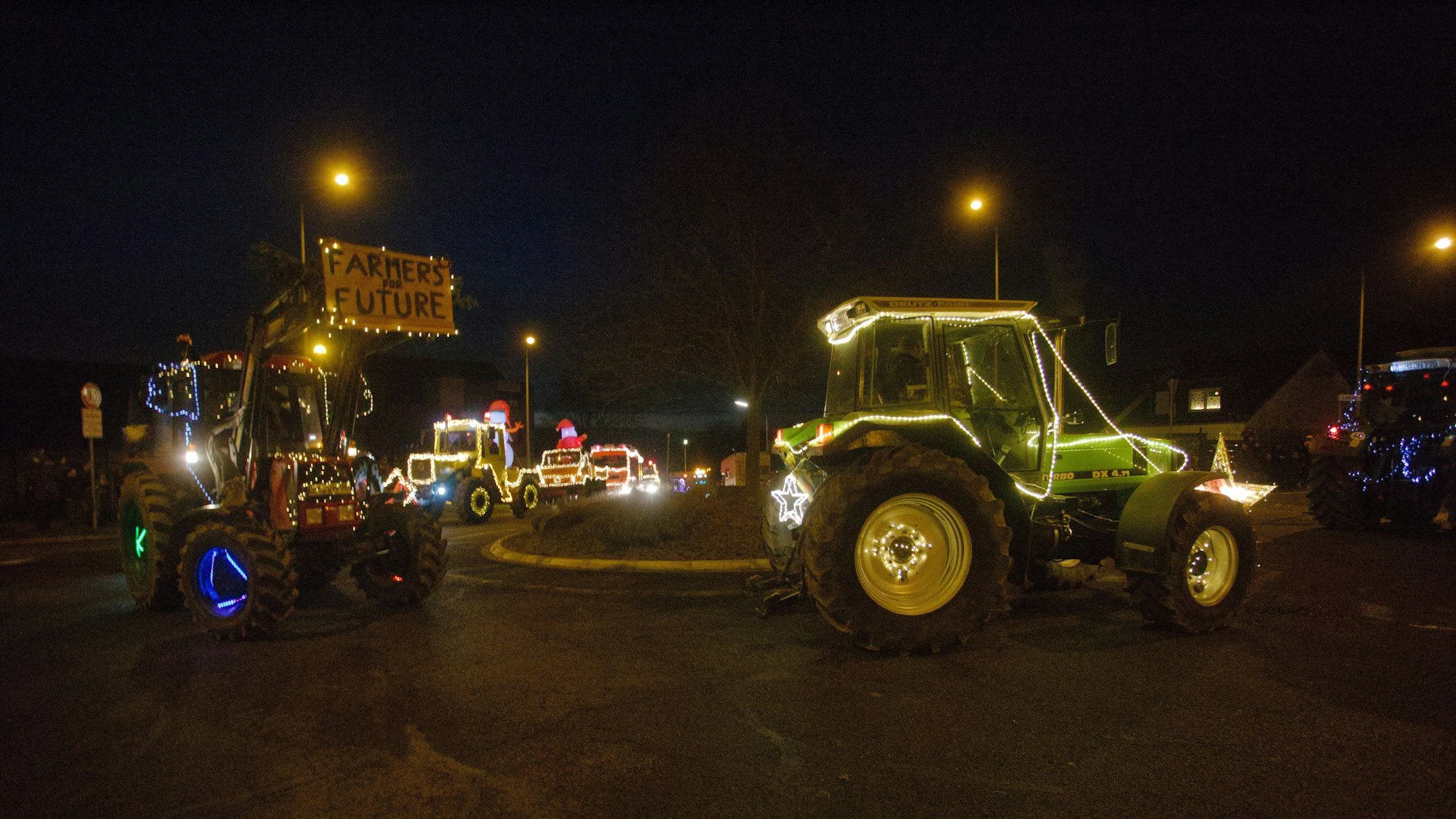 Mit bunten Lichtern geschmückte Traktoren fahren durch einen Kreisverkehr bei Nacht.