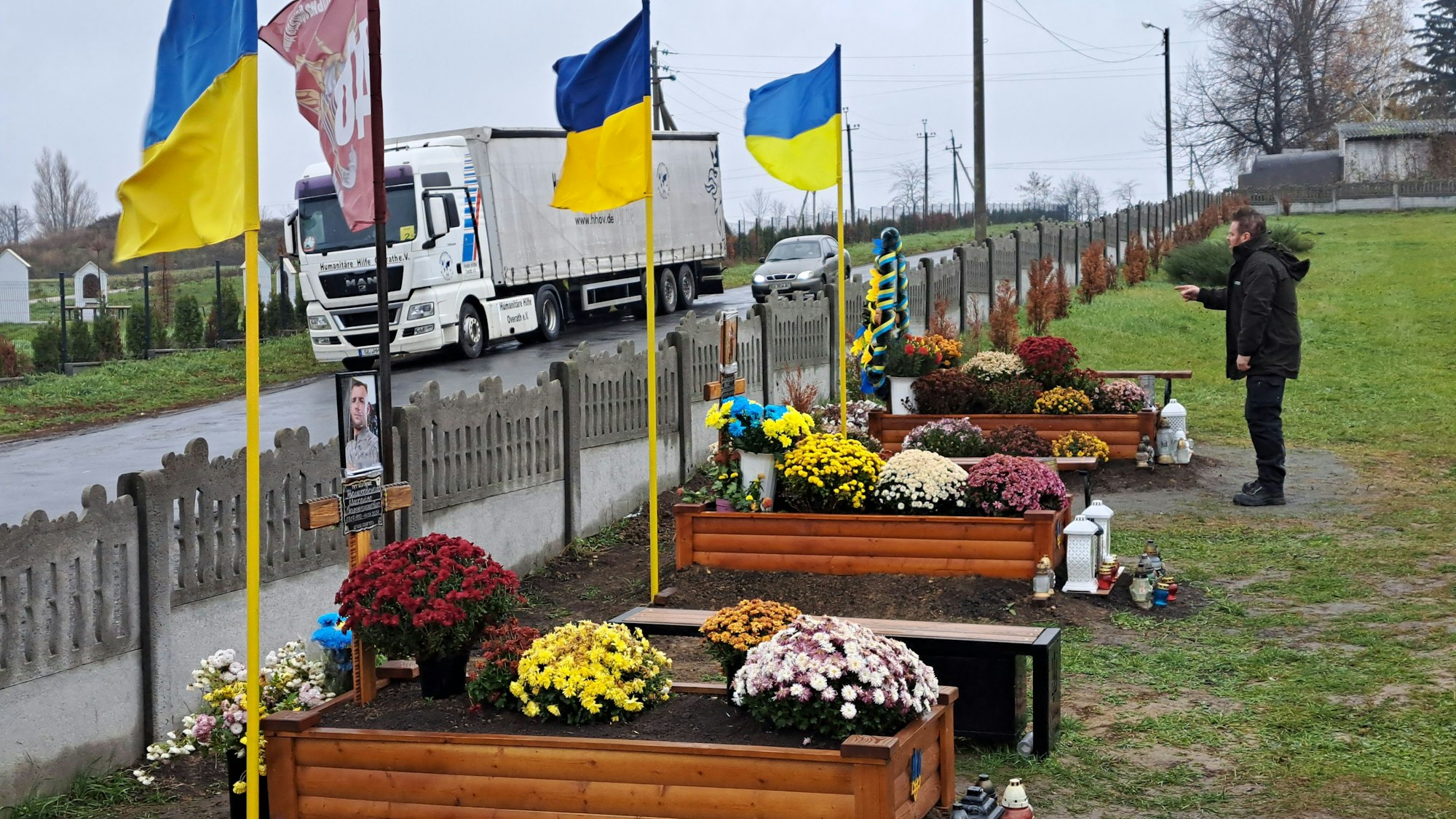 Ein Mann steht vor Gräbern mit ukrainischen Fahnen, im Hintergrund ist ein Lkw der Humanitären Hilfe Overath zu sehen.