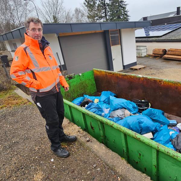 Der Bauhofleiterin orangefarbener Warnjacke steht neben einem grünen Container voller Müllsäcke.