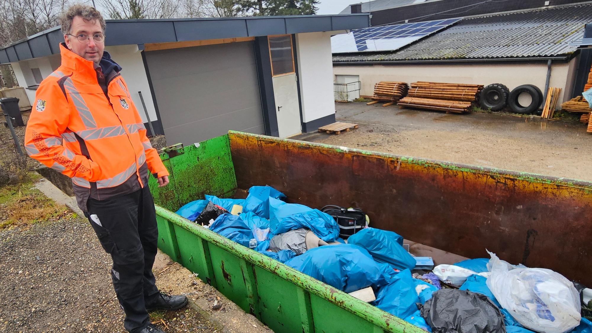 Der Bauhofleiterin orangefarbener Warnjacke steht neben einem grünen Container voller Müllsäcke.