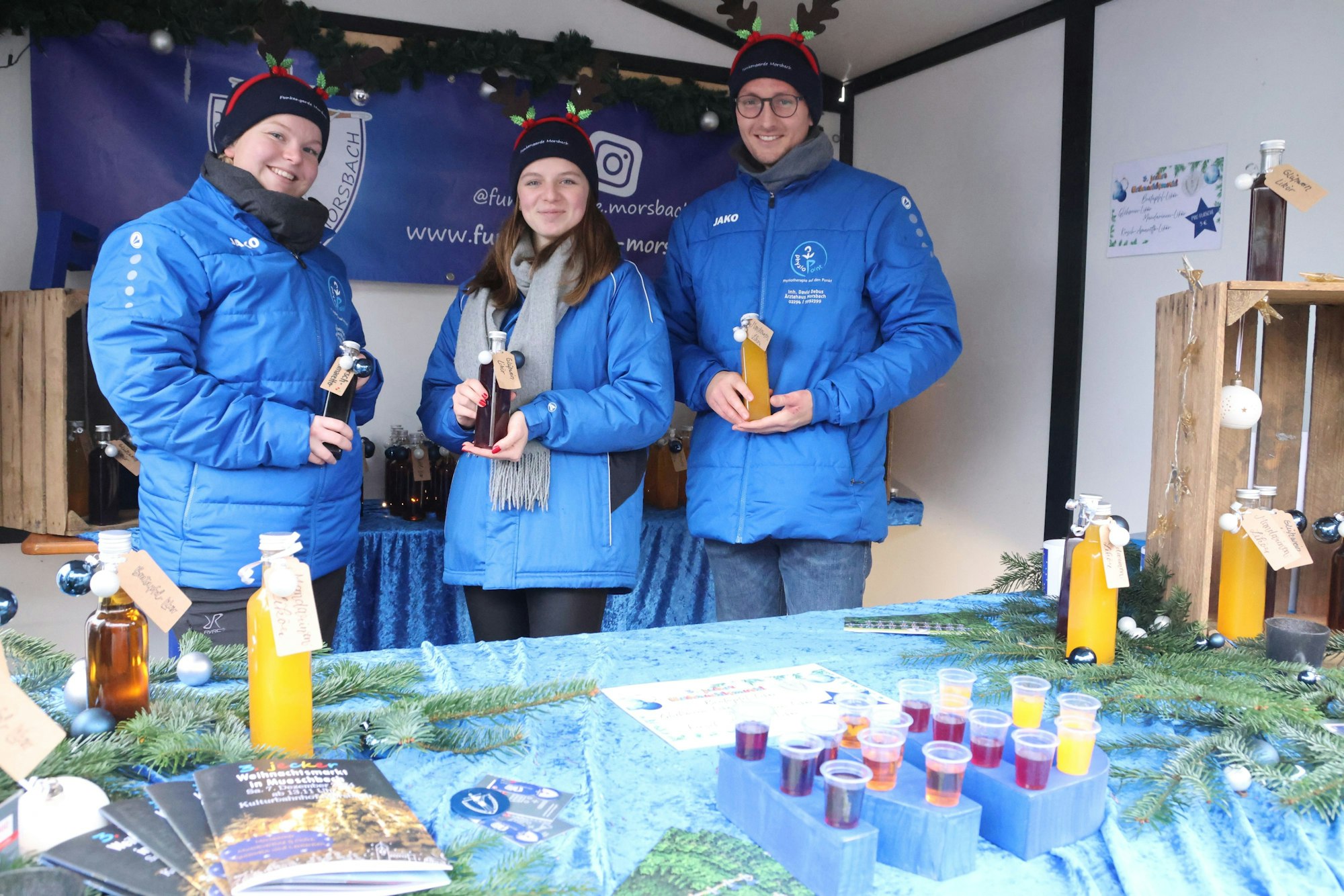 Zwei Frauen und ein Mann stehen hinter einem Verkaufstisch und haben Flaschen in Händen, sie tragen blaue Jacken.