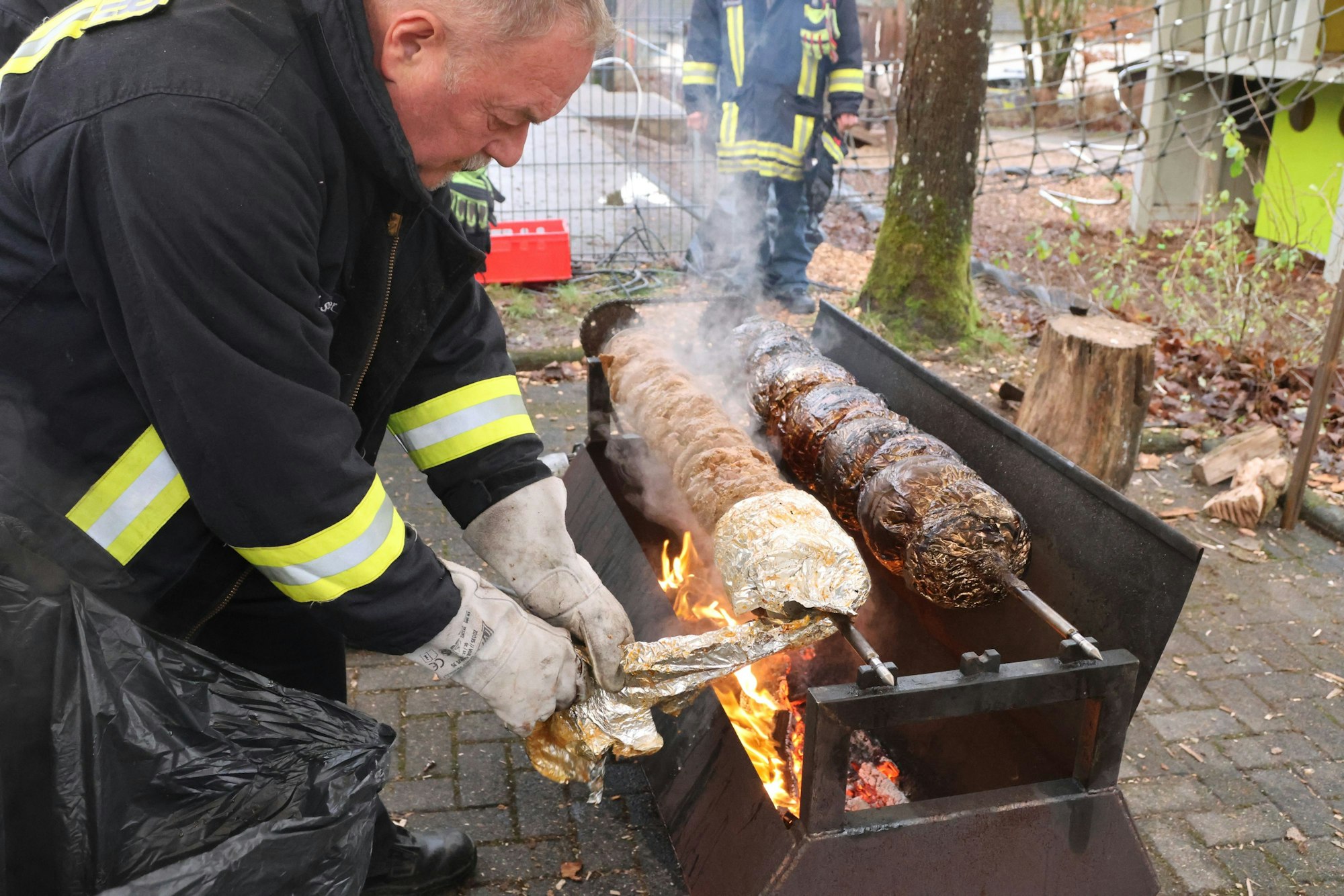 Heinz-Josef Stangier trägt eine schwarze Feuerwehrjacke und dicke Handschuhe und befreit den Spießbraten von seiner Umhüllung aus Alu-Folie. Darunter prasseln Flammen auf einem Grill.