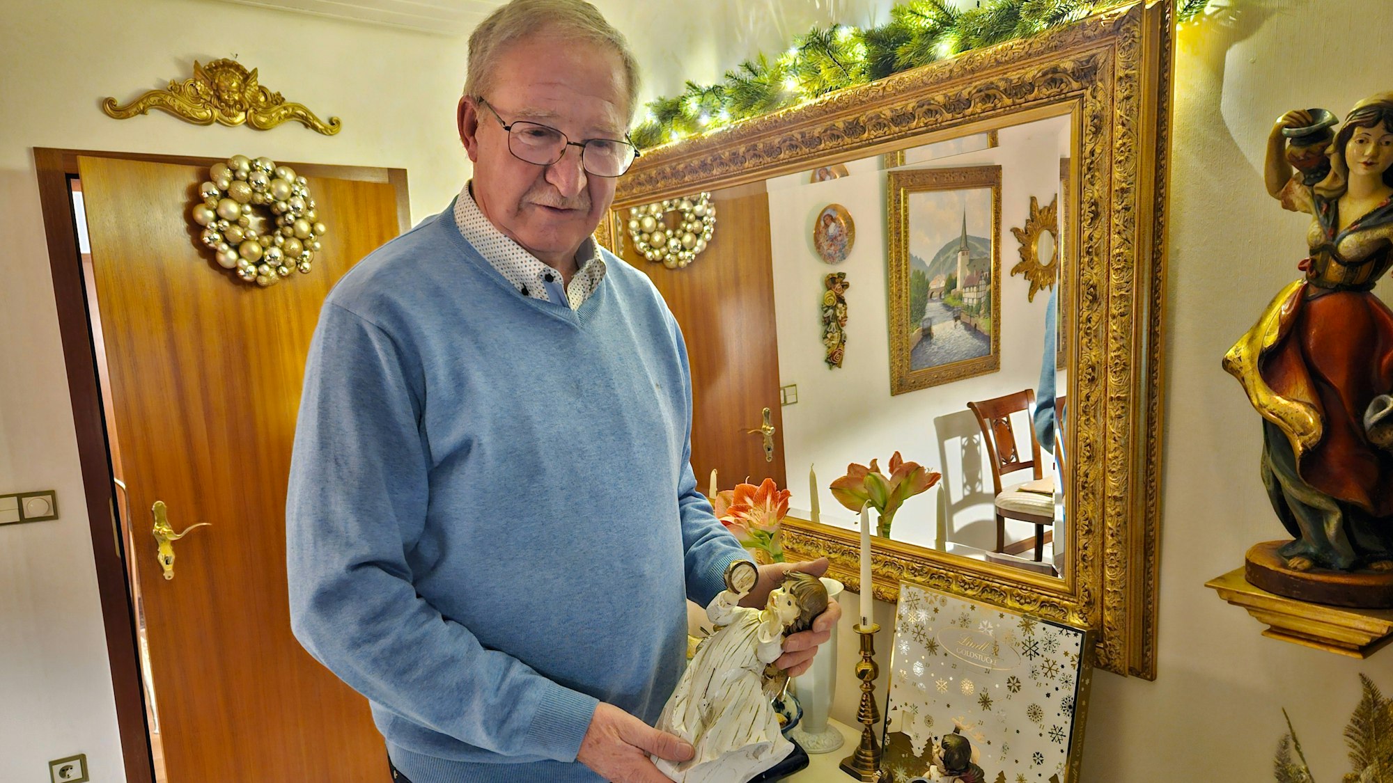 Georg Weber steht im Wohnzimmer vor einem Spiegel und hält eine Engelsfigur.
