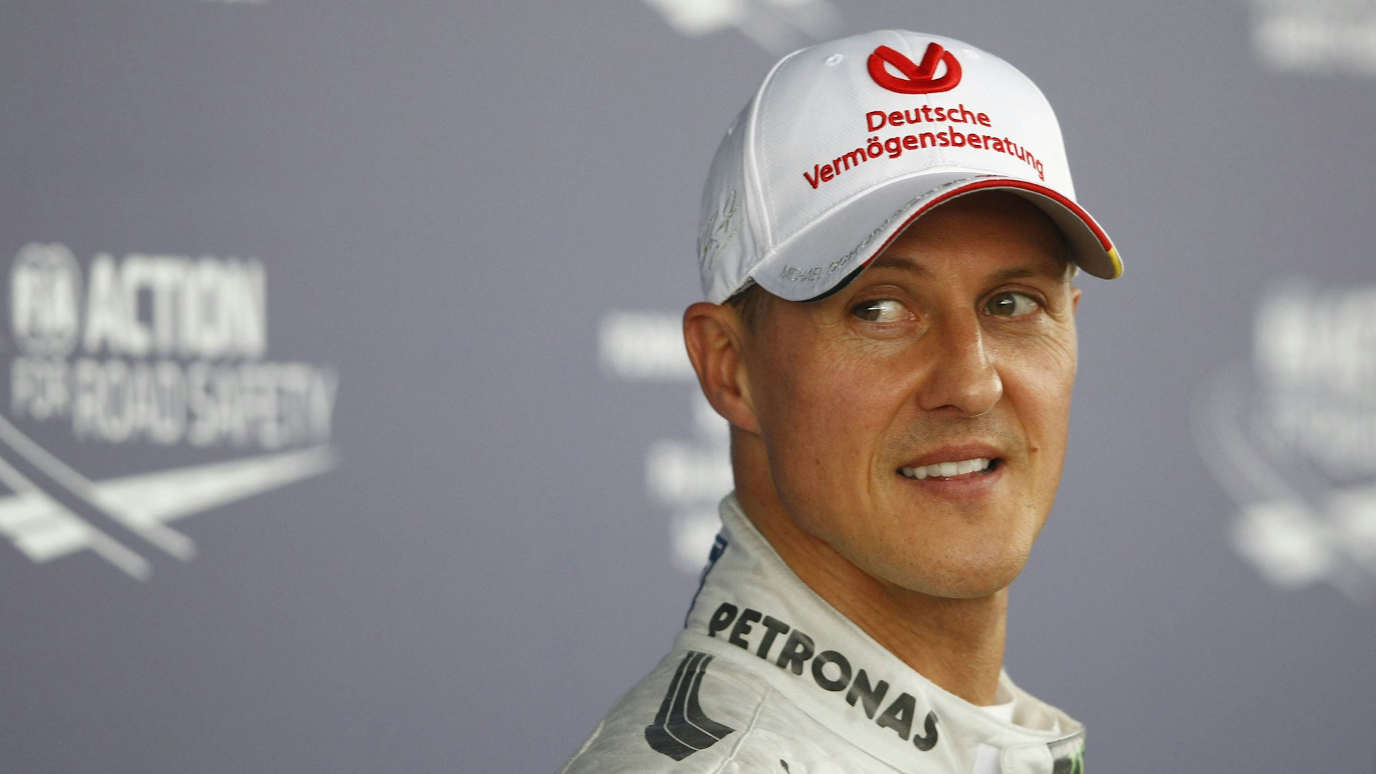 Michael Schumacher ist im Porträt zu sehen. Er trägt eine Baseballkappe und lächelt. Sein Blick ist an der Kamera vorbei gerichtet.