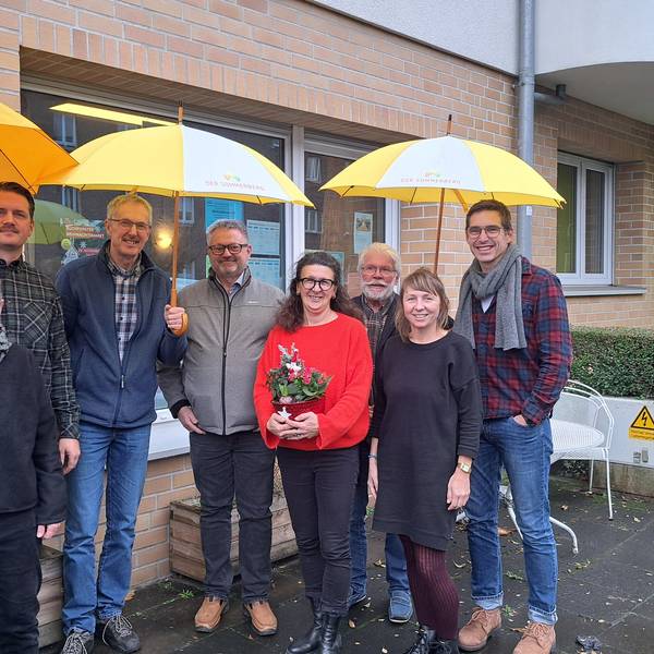 Acht Menschen stehen vor einem Backsteingebäude. Drei von ihnen halten gelbe Regenschirme.