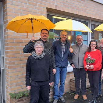 Acht Menschen stehen vor einem Backsteingebäude. Drei von ihnen halten gelbe Regenschirme.