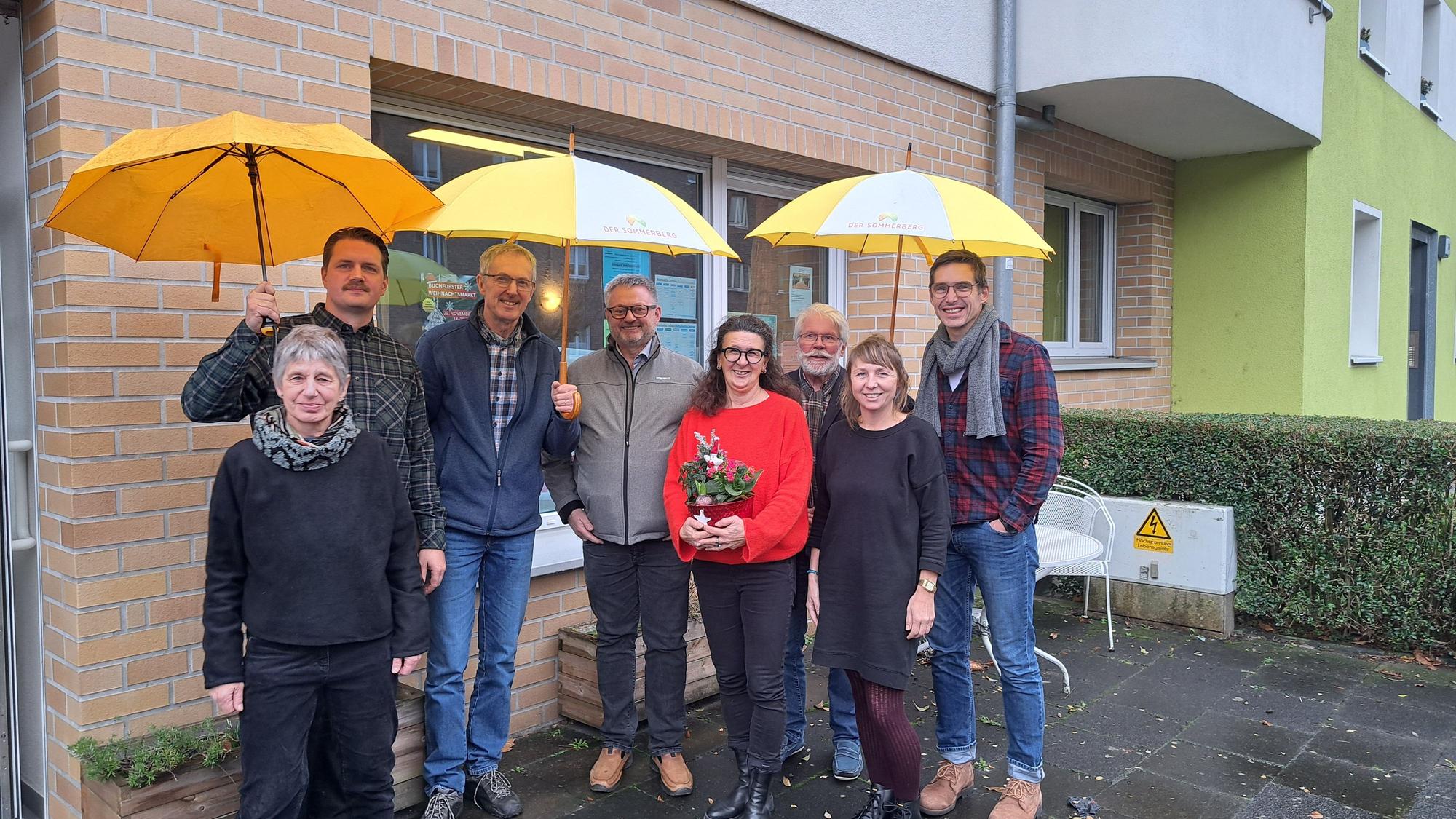 Acht Menschen stehen vor einem Backsteingebäude. Drei von ihnen halten gelbe Regenschirme.