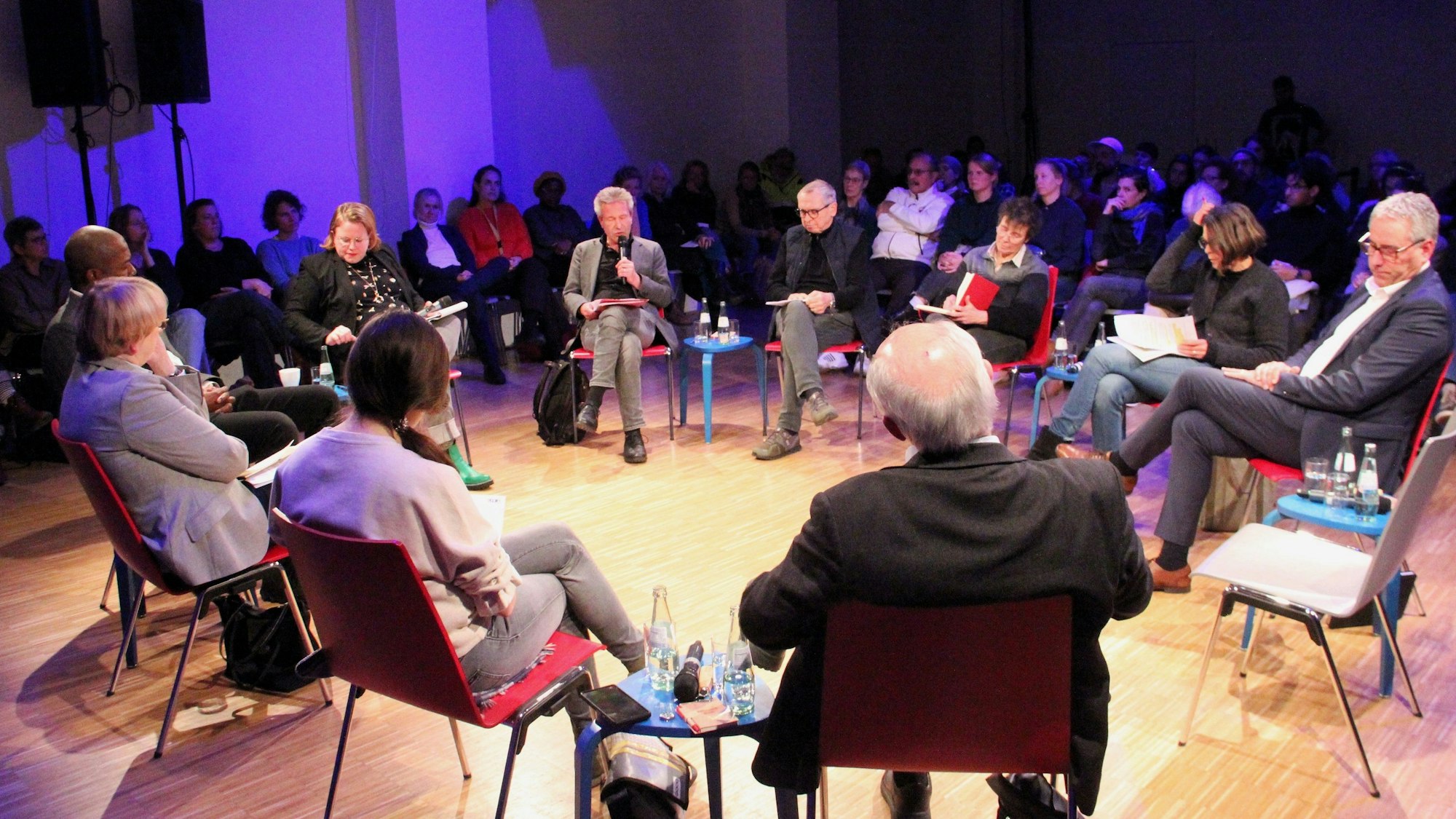 Zehn Menschen sitzen bei einer Podiumsdiskussion in einem Stuhlkreis. Im Hintergrund sitzt Publikum.